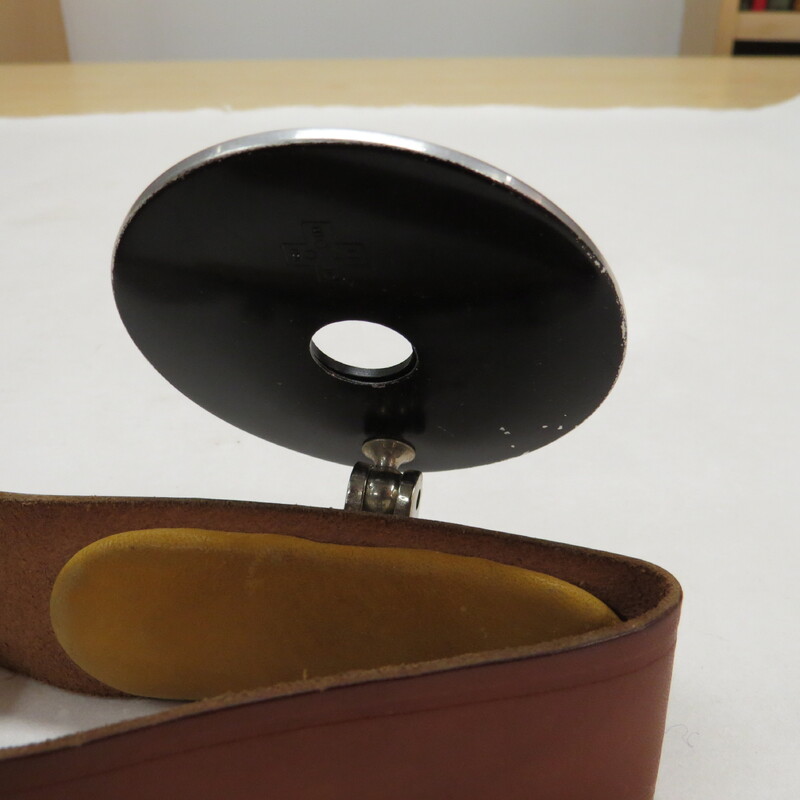 Adjustable head mirror with adjustable leather strap. Mirror is circular, with a circular aperture and a slightly convex lens encased and backed by steel, which is attached by a fully rotatable and adjustable screw mechanism which allows the mirror to be completely detached from its harness. Strap is of thick brown leather, and has a steel clasp which a snap mechanism for adjusting its length.