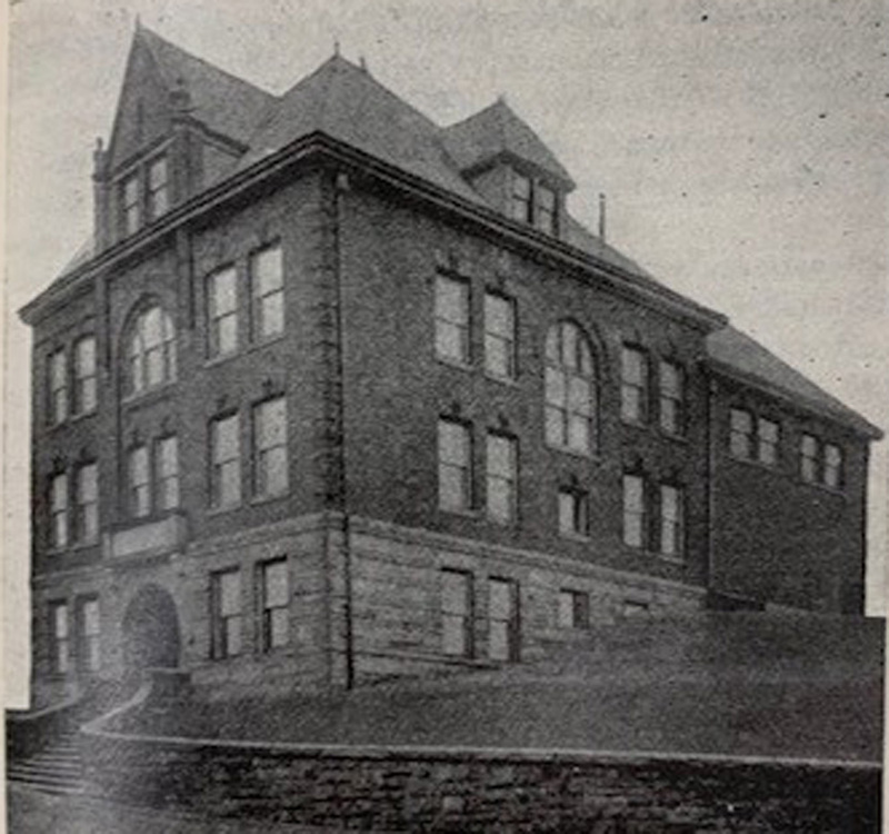 A black-and-white, grainy photograph of a square three-story brick building with dormers.