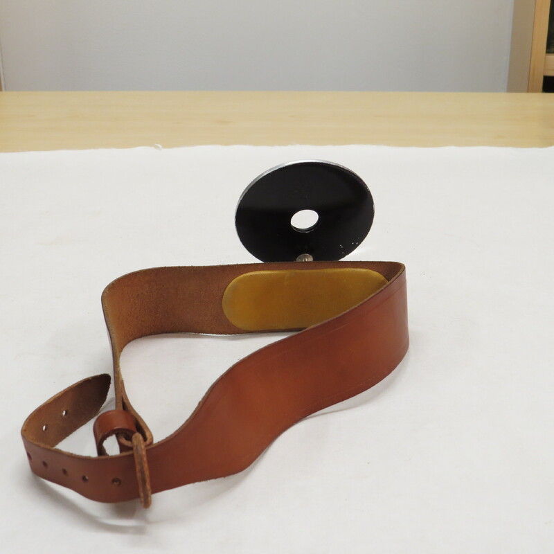 Adjustable head mirror with adjustable leather strap. Mirror is circular, with a circular aperture and a slightly convex lens encased and backed by steel, which is attached by a fully rotatable and adjustable screw mechanism which allows the mirror to be completely detached from its harness. Strap is of thick brown leather, and has a steel clasp which a snap mechanism for adjusting its length.