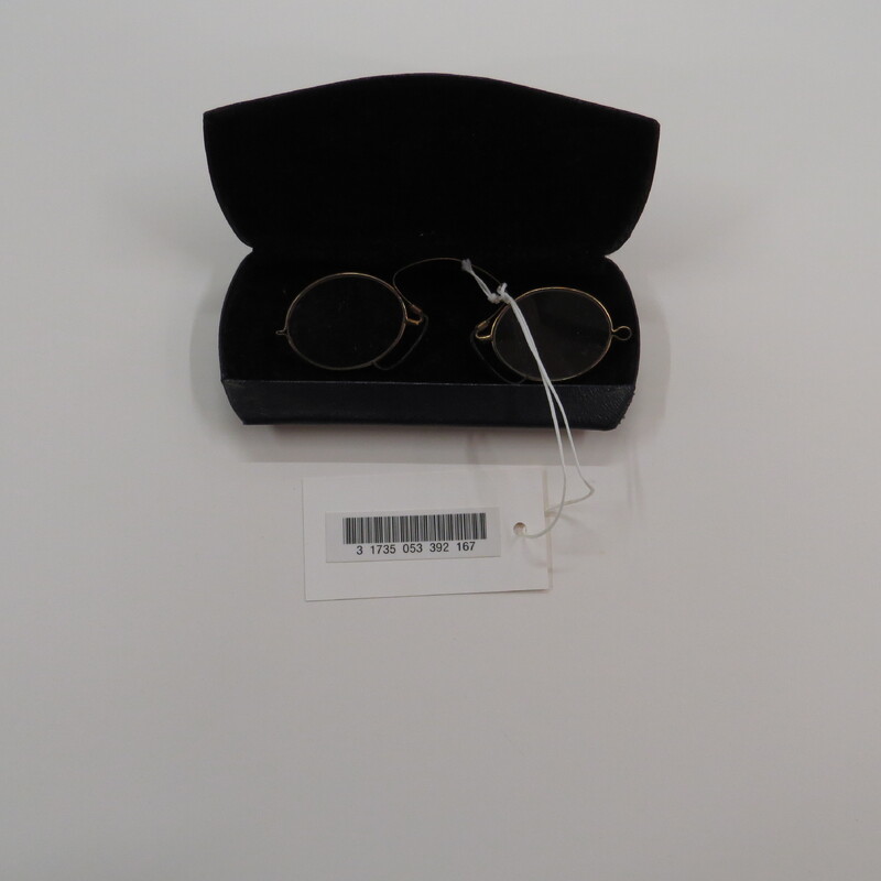 Oval lenses in gold tone frames with a spring clip and black pads in a clamshell metal case covered with leather-like material.