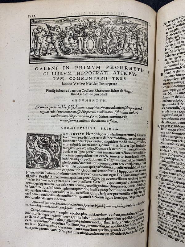 A photo of an open old book, showing a illustration of angels at the top of the page and Latin text.
