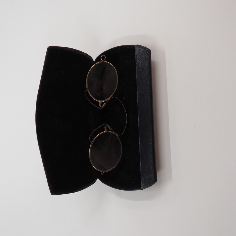 Oval lenses in gold tone frames with a spring clip and black pads in a clamshell metal case covered with leather-like material.