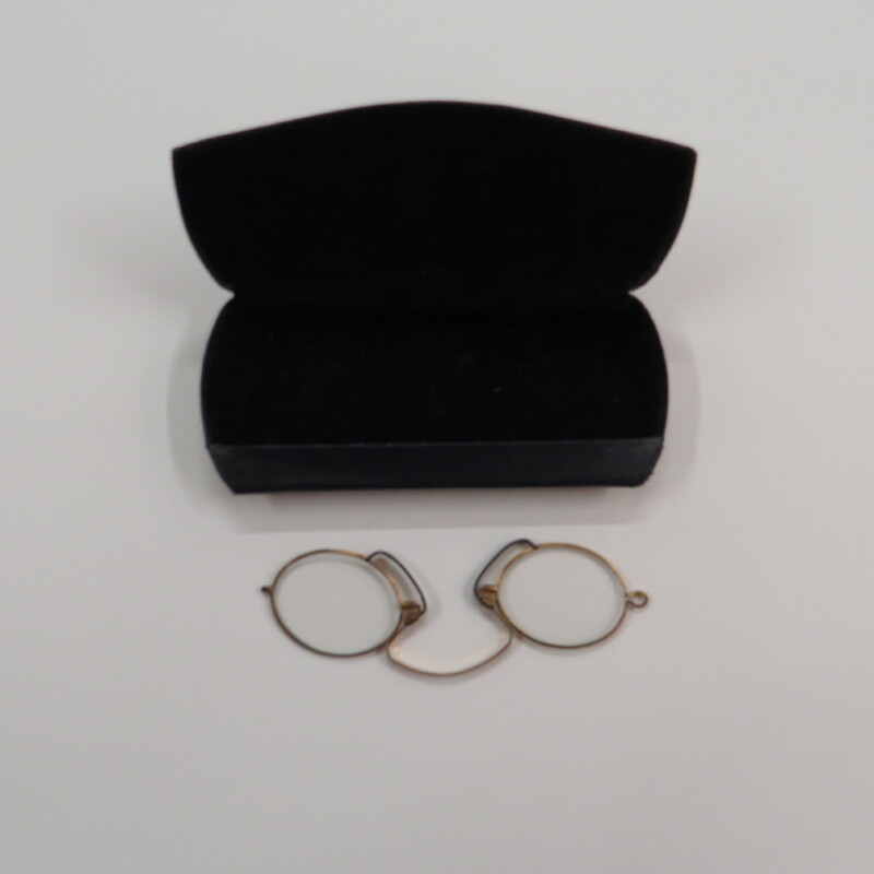 Oval lenses in gold tone frames with a spring clip and black pads in a clamshell metal case covered with leather-like material.