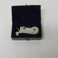 A white metal spring lancet with a button trigger in a paper box lined with velvet, closed by latch.