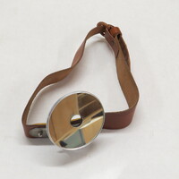 Adjustable head mirror with adjustable leather strap. Mirror is circular, with a circular aperture and a slightly convex lens encased and backed by steel, which is attached by a fully rotatable and adjustable screw mechanism which allows the mirror to be completely detached from its harness. Strap is of thick brown leather, and has a steel clasp which a snap mechanism for adjusting its length.